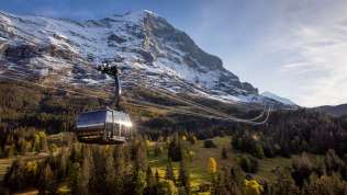 The Eiger Express cable car in spring, with green countryside in the foreground, snow-capped mountains in the background, and a blue sky.